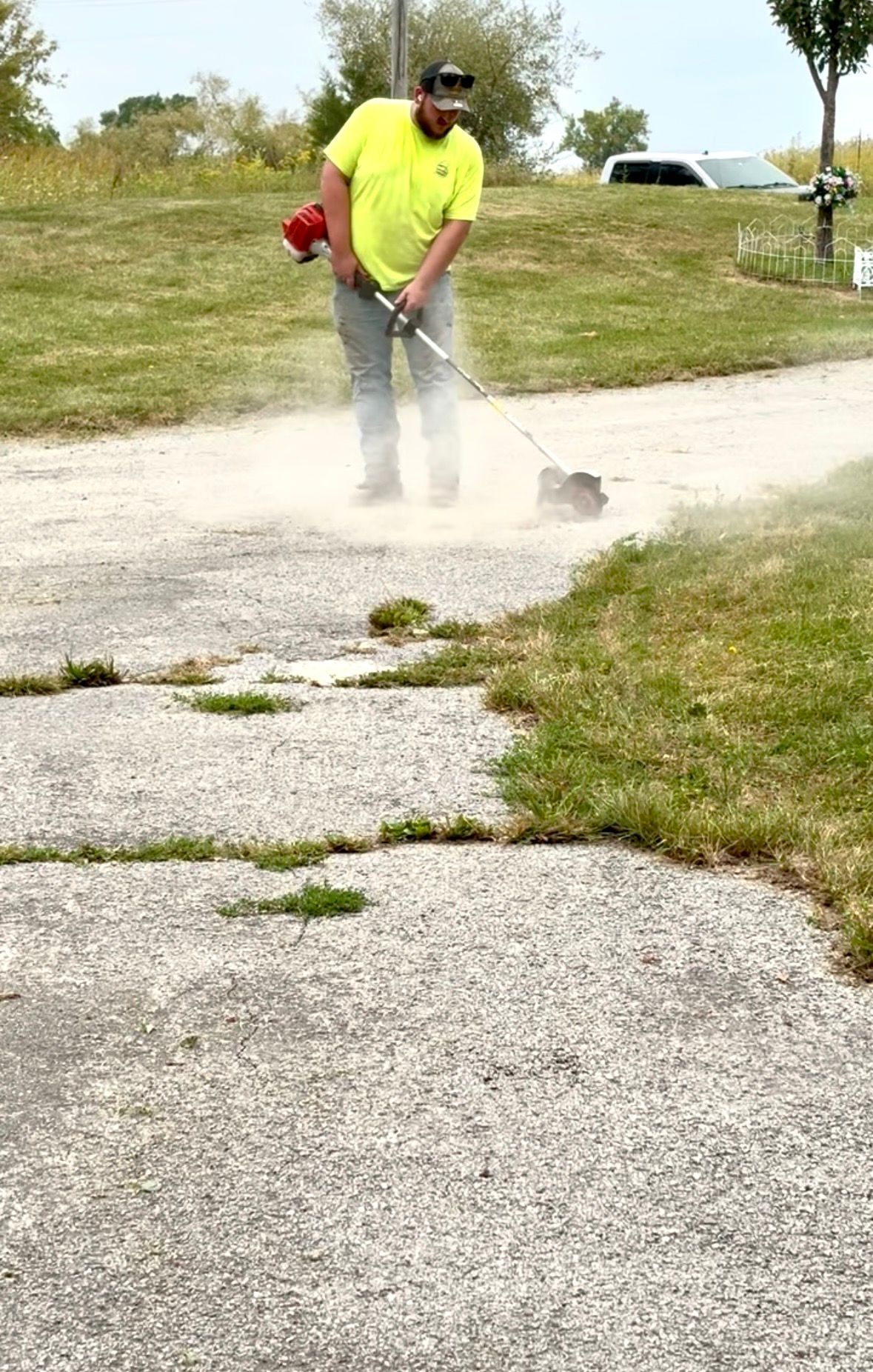 Worker in neon yellow shirt using a weed trimmer on a gravel path beside a grassy area.