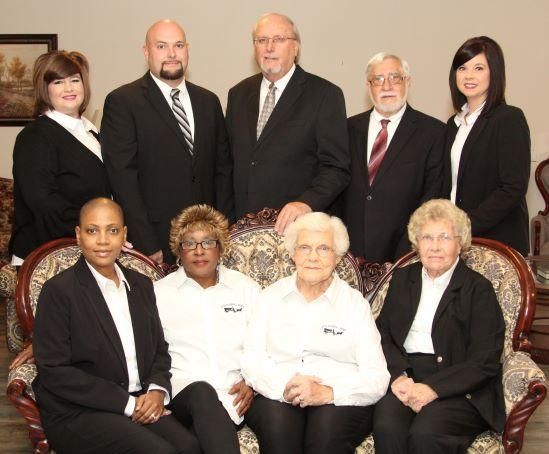 Group portrait of eight people in formal attire posed indoors on chairs and standing behind them