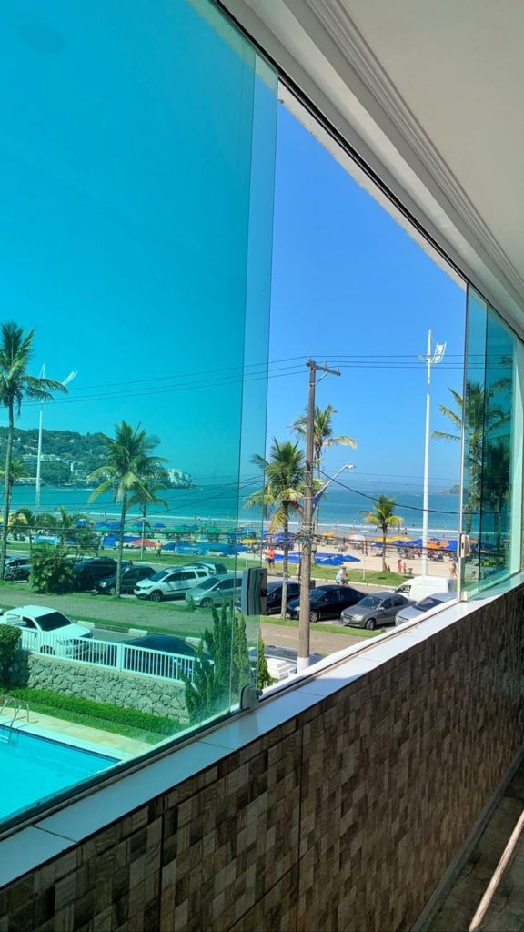 Vista da varanda para uma praia ensolarada, oceano, palmeiras, carros estacionados e um parapeito de vidro azulado.