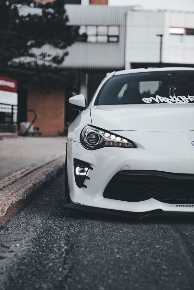 Carro esportivo branco estacionado na rua, vista frontal em close-up com fundo urbano desfocado.