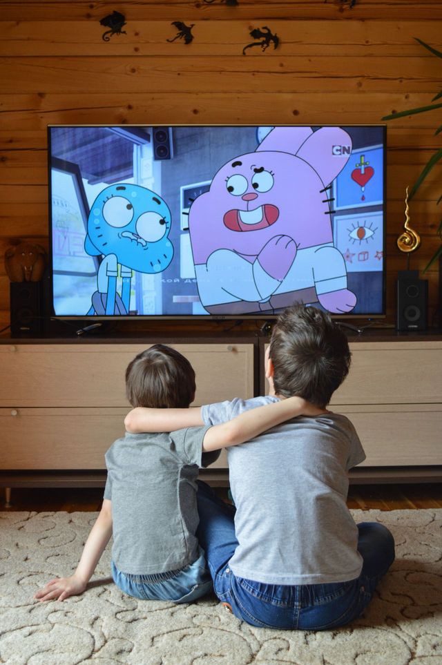 Duas crianças sentam-se no chão assistindo a um desenho animado na TV, em uma sala aconchegante com painéis de madeira.