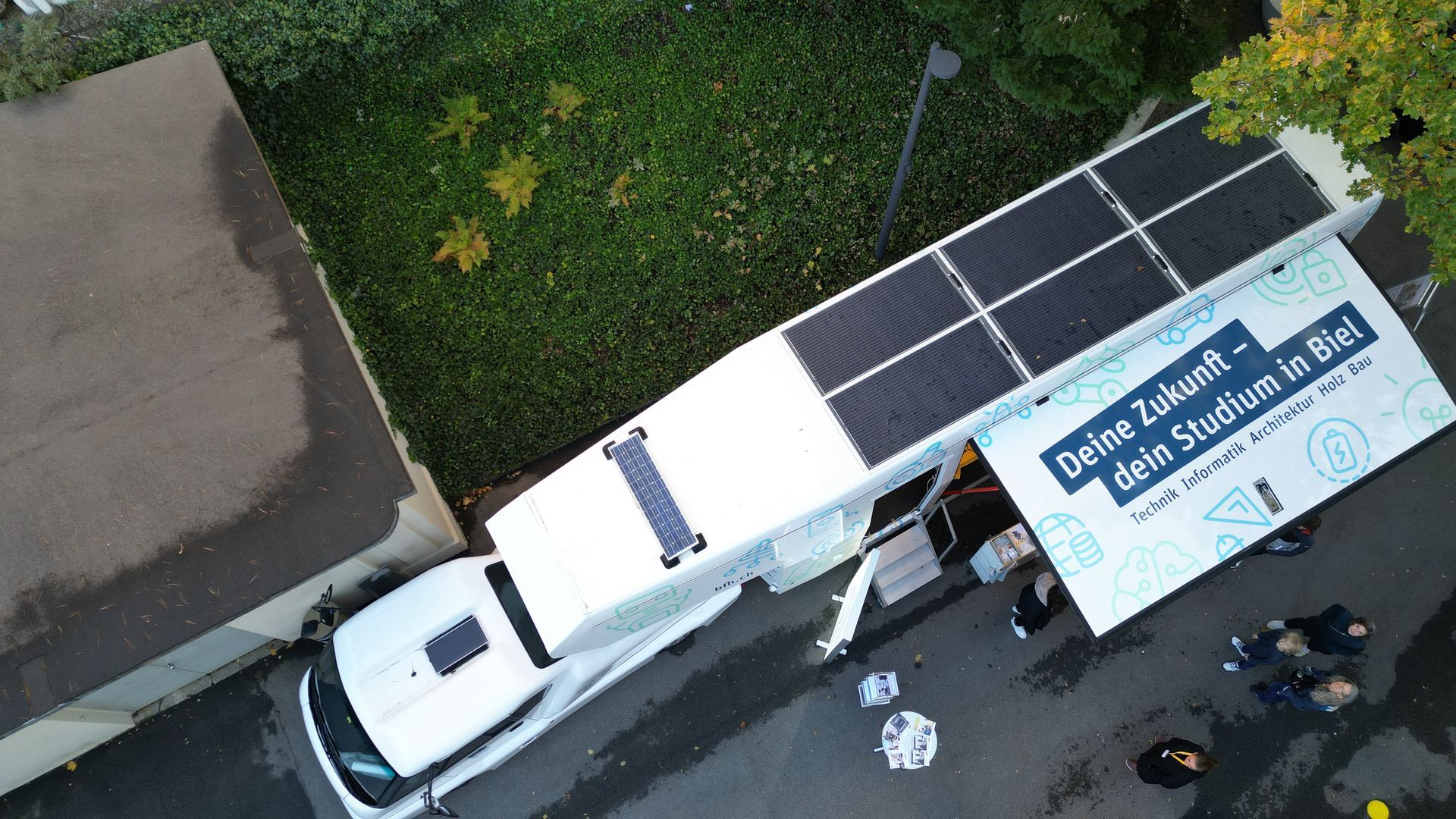 Luftaufnahme eines weißen Werbe-Lkw mit Solarpaneelen, der im Freien geparkt ist; Menschen stehen in der Nähe der Markise.