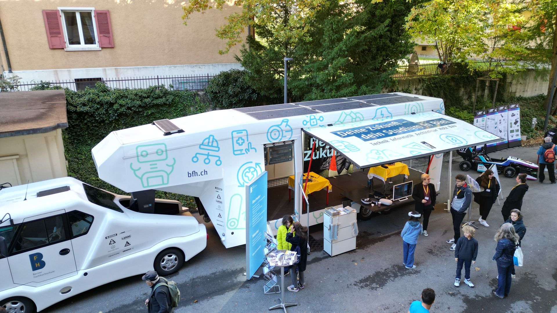 Ein mobiles Technologie-Klassenzimmer im Freien mit weißer Außenhülle und blauen Symbolen steht in einem Innenhof und ist von neugierigen Besuchern umringt.