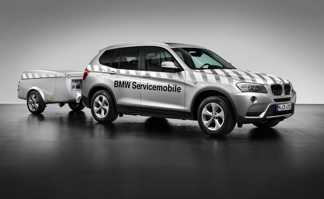 Ein silberner BMW Servicemobile SUV mit karierter Lackierung zieht einen passenden Anhänger in einer Studio-Kulisse.