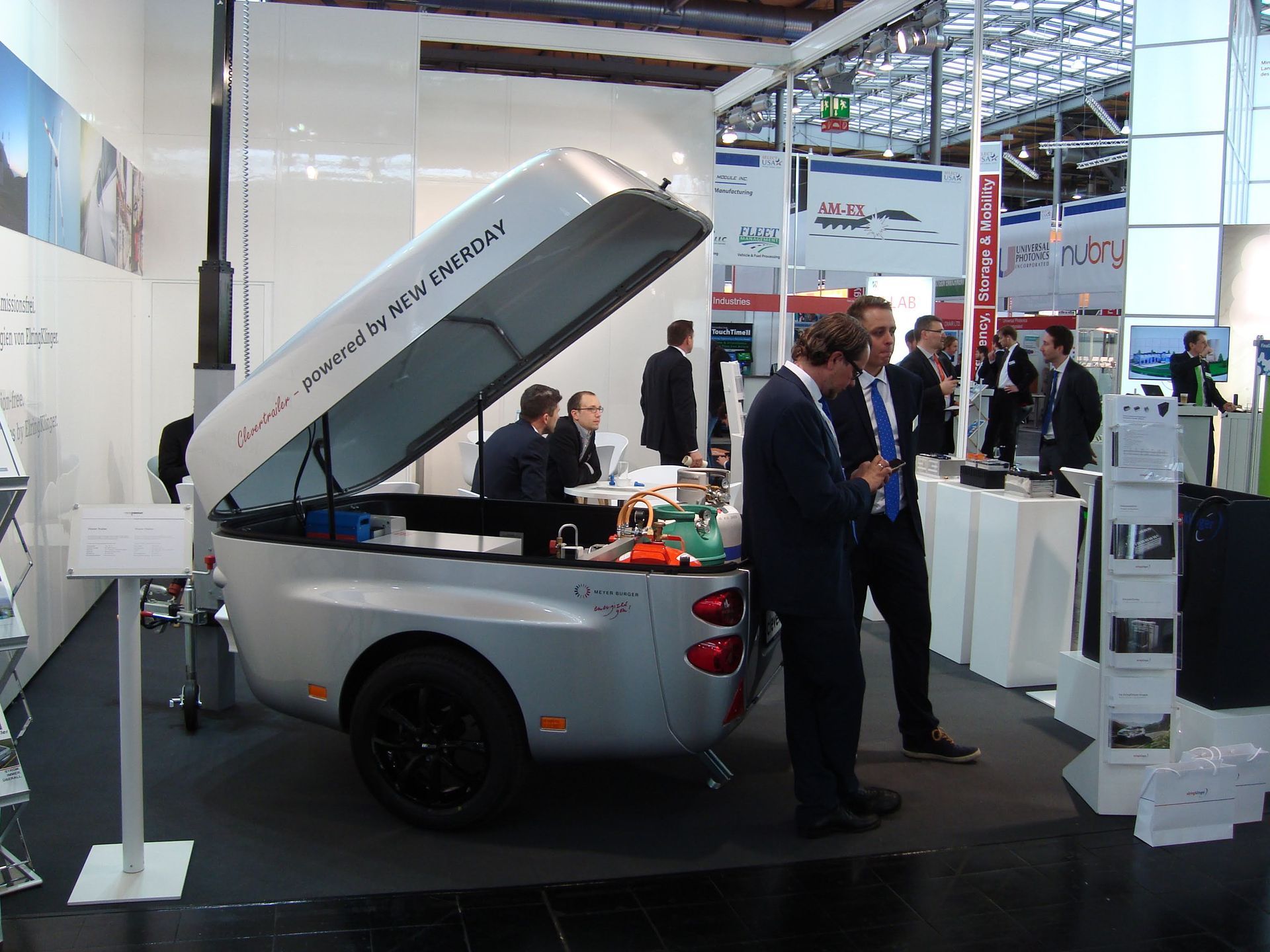 Ein silberner, tropfenförmiger Wohnwagen mit geöffnetem Deckel, ausgestellt auf einer Messe; zwei Personen in Anzügen stehen daneben.