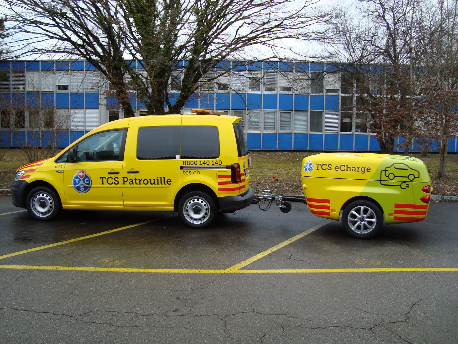 Ein gelber TCS Patrol-Van zieht einen passenden mobilen Anhänger mit Ladestation für Elektrofahrzeuge auf einem Parkplatz im Freien.