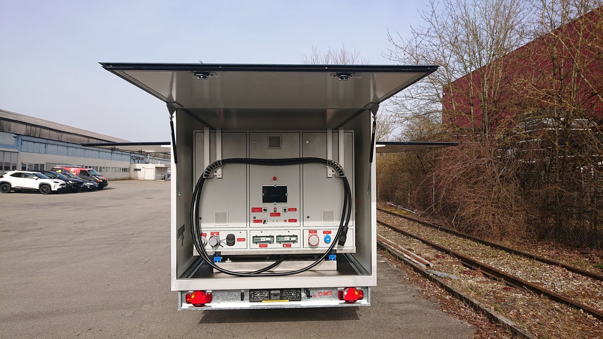 Ein im Freien geparkter Anhänger mit geöffneter Seitenluke gibt den Blick auf ein großes elektrisches Industriegerät im Inneren frei.