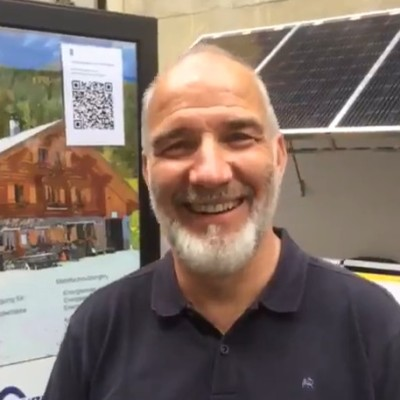 Eine lächelnde Person steht vor einem Display mit einer Hausillustration, einem QR-Code und Solarpaneelen.