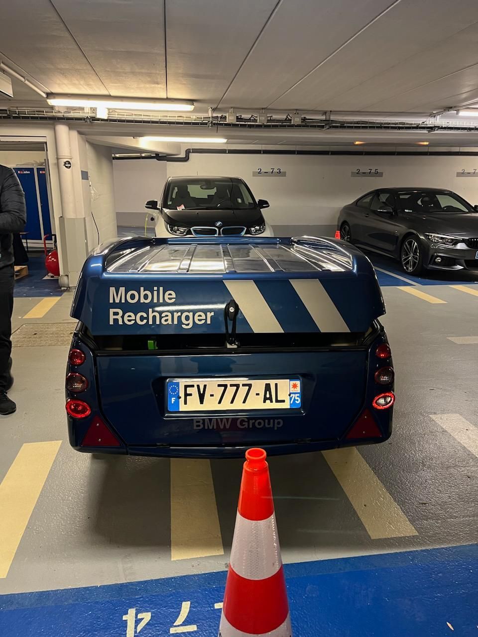 Ein blauer BMW Group „Mobile Recharger“-Serviceanhänger parkte in einer Tiefgarage hinter einem orangefarbenen Verkehrskegel.