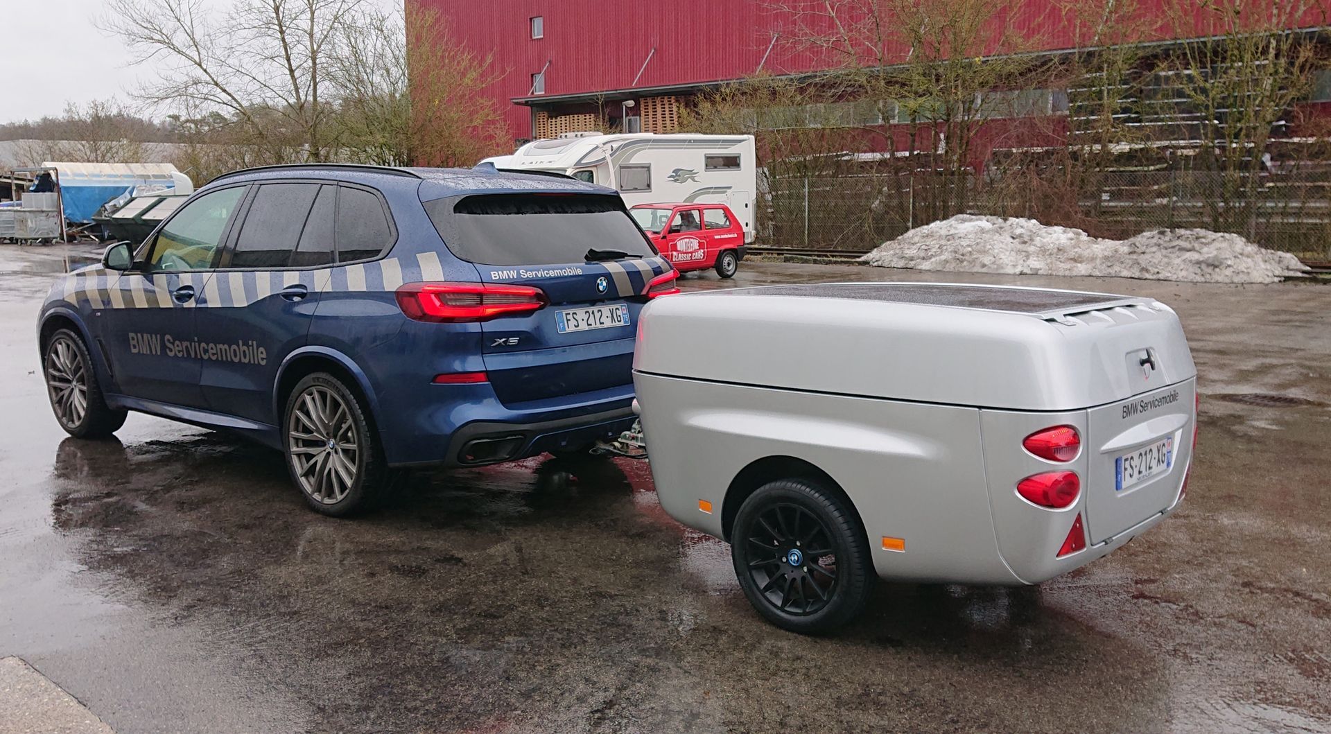 Ein blauer BMW SUV zieht einen kompakten, silbernen, aerodynamischen Anhänger auf einem Parkplatz im Freien.