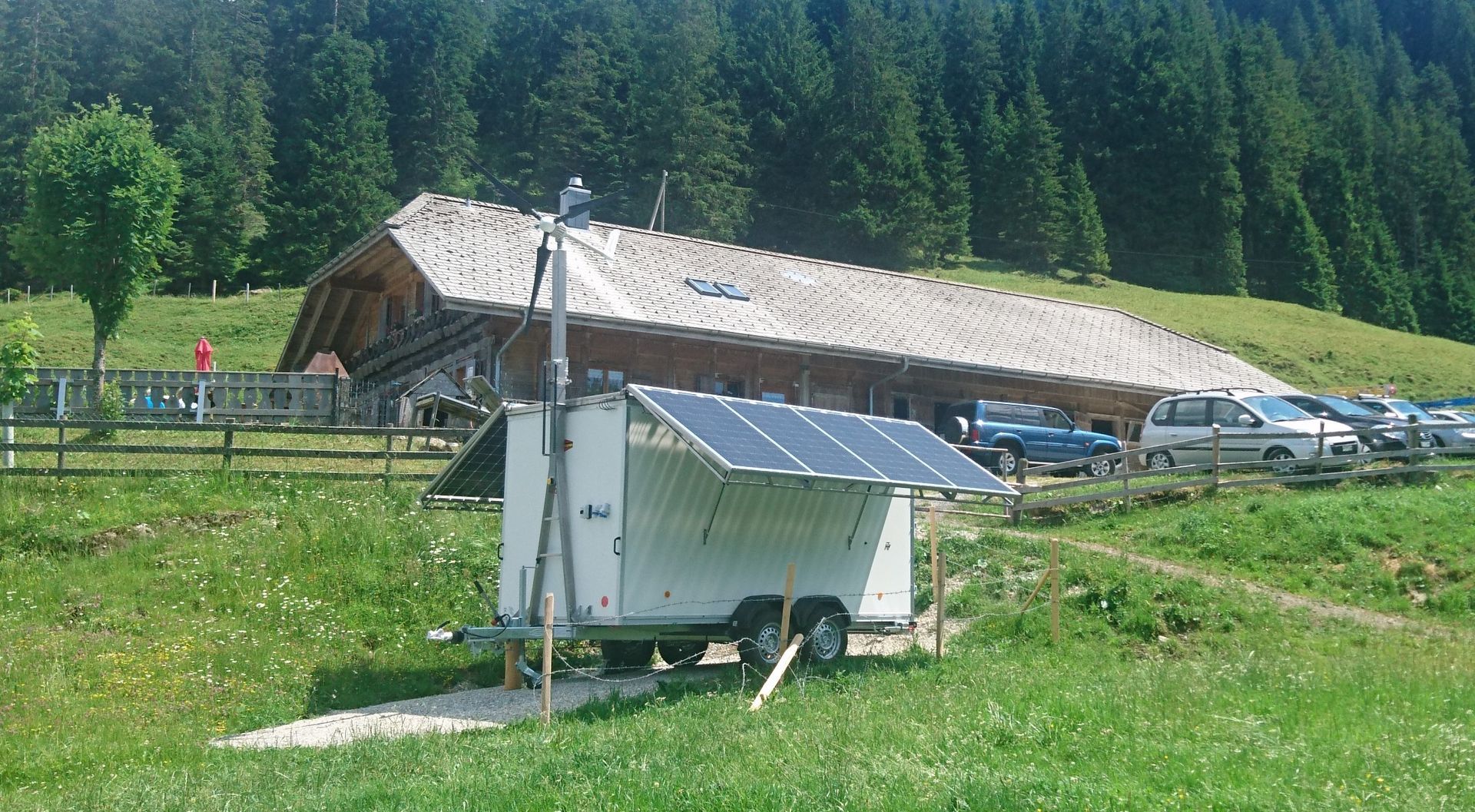 Ein mobiler Anhänger mit auf dem Dach installierten Solarpaneelen steht auf einer grasbewachsenen Wiese vor einem rustikalen Holzgebäude.