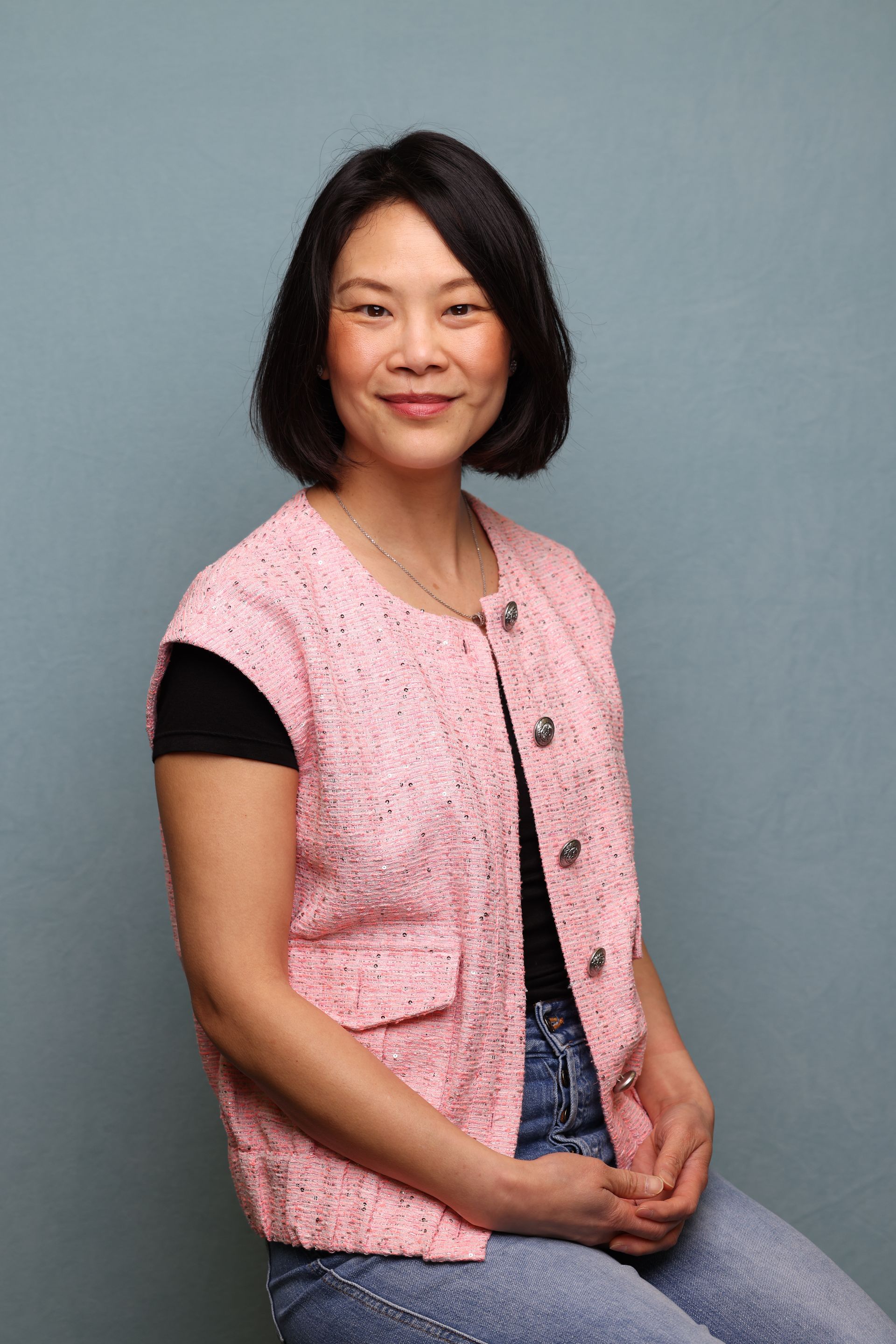 A woman in a pink vest and jeans is sitting in front of a blue wall.