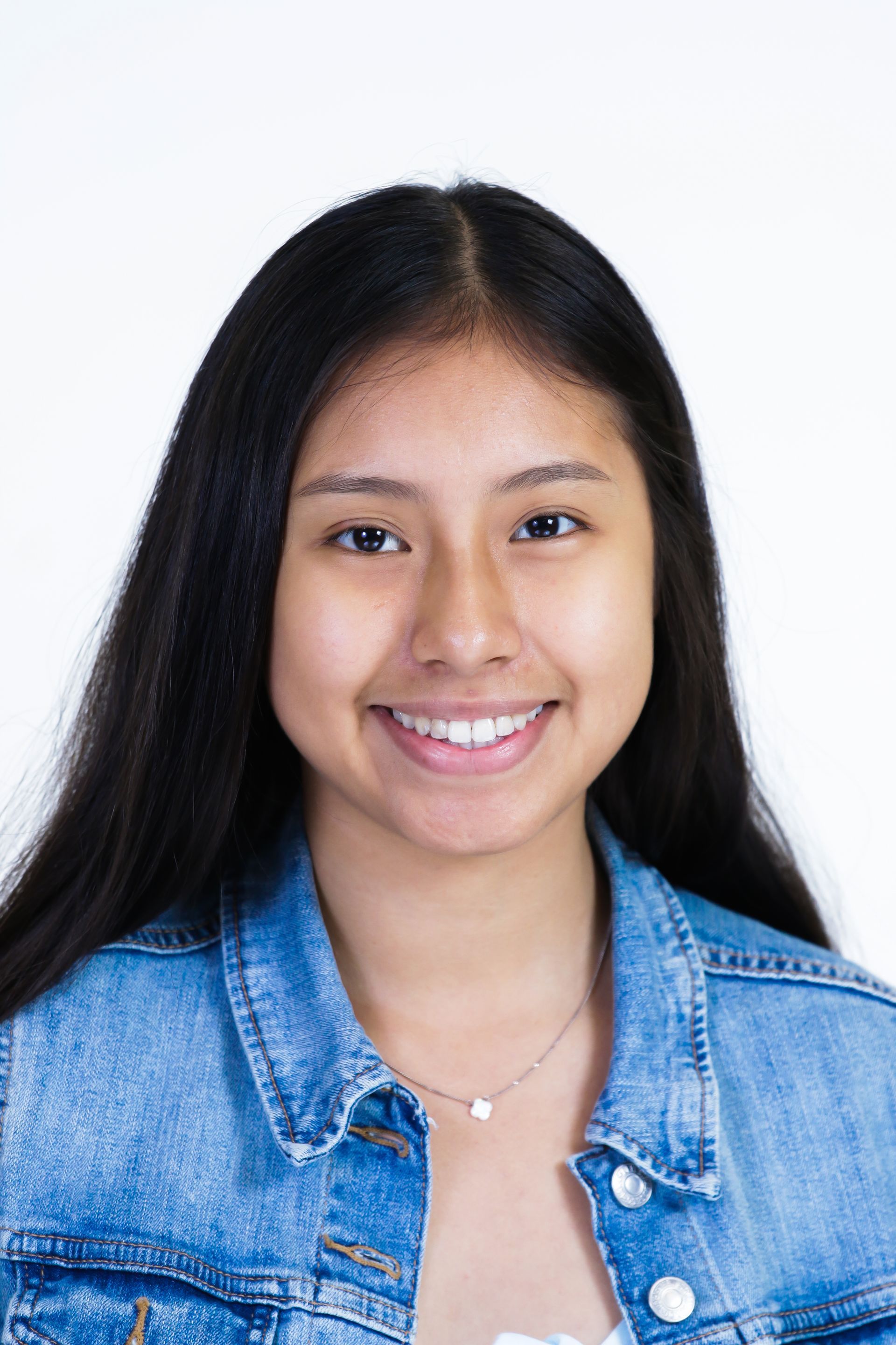 A young woman wearing a denim jacket and a necklace is smiling for the camera.