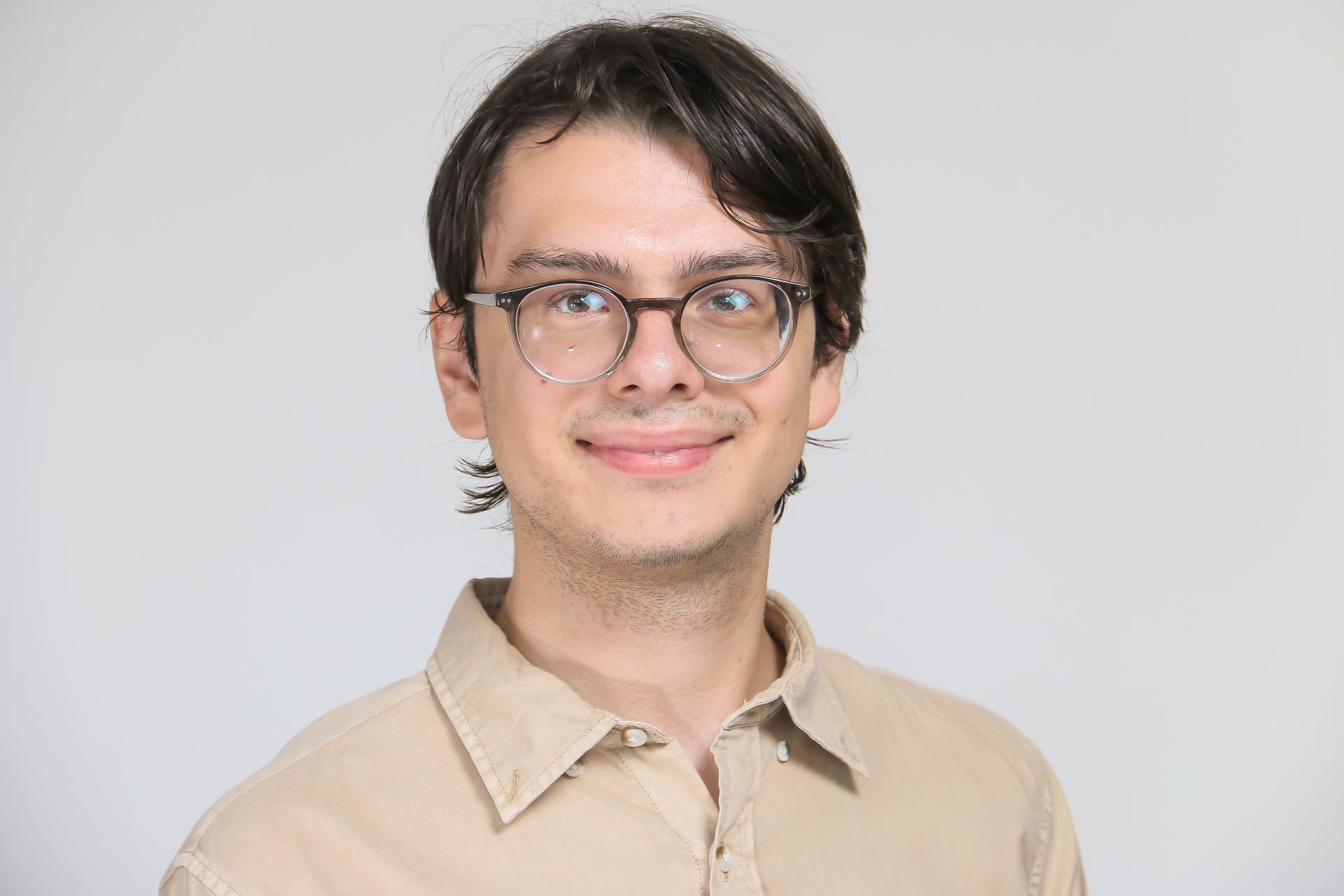 A young man wearing glasses and a tan shirt is smiling for the camera.