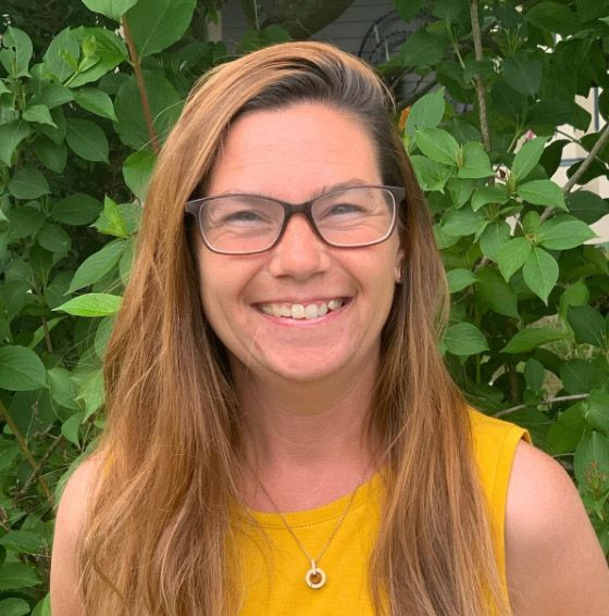 A woman wearing glasses and a yellow shirt is smiling in front of a bush.