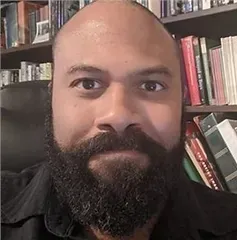 A bald man with a beard is sitting in front of a bookshelf.