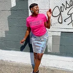 A woman in a pink shirt and silver skirt is standing in front of a wall.