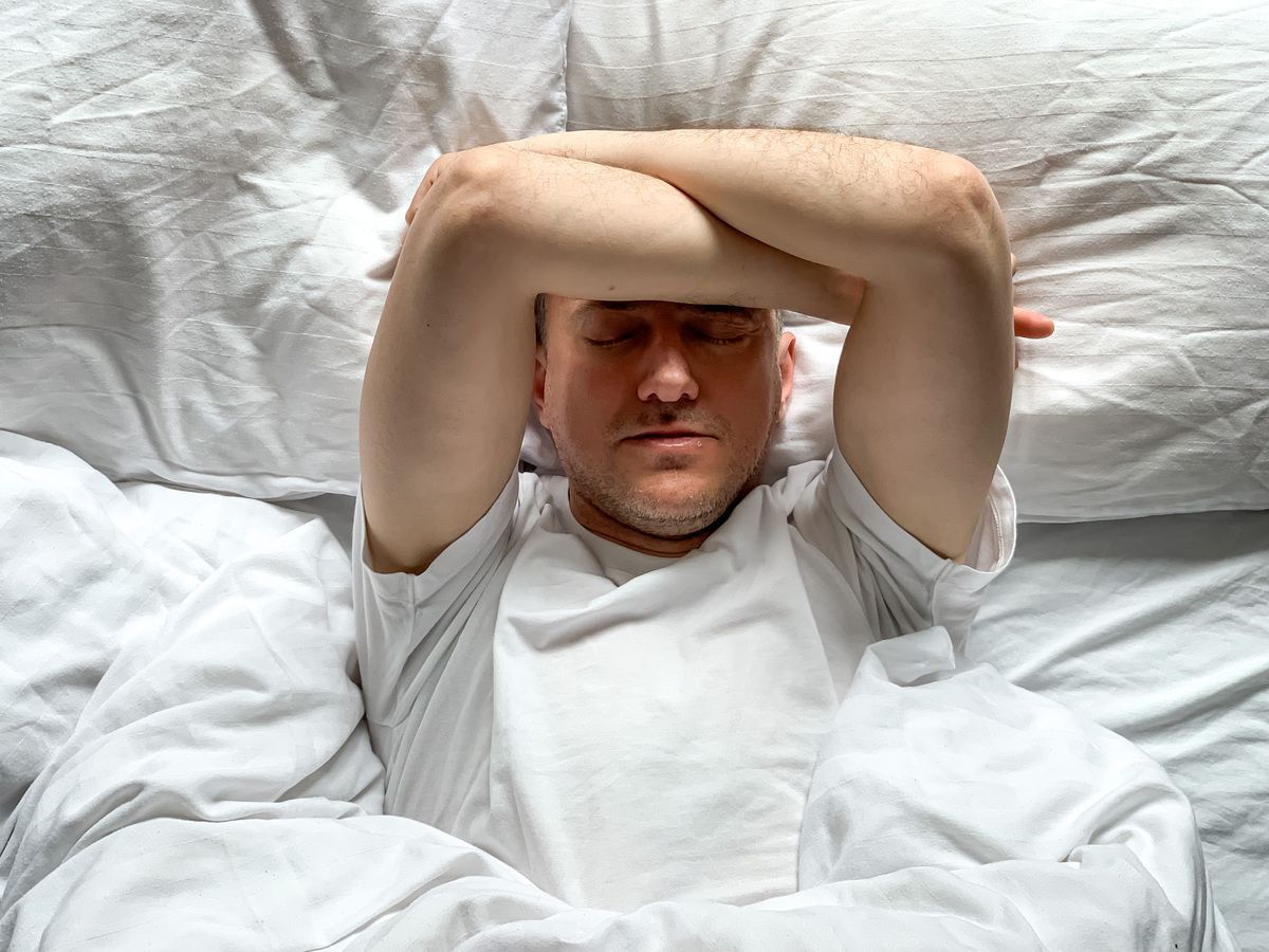 Person lying in bed with eyes closed, arms resting above their head on white bedding.