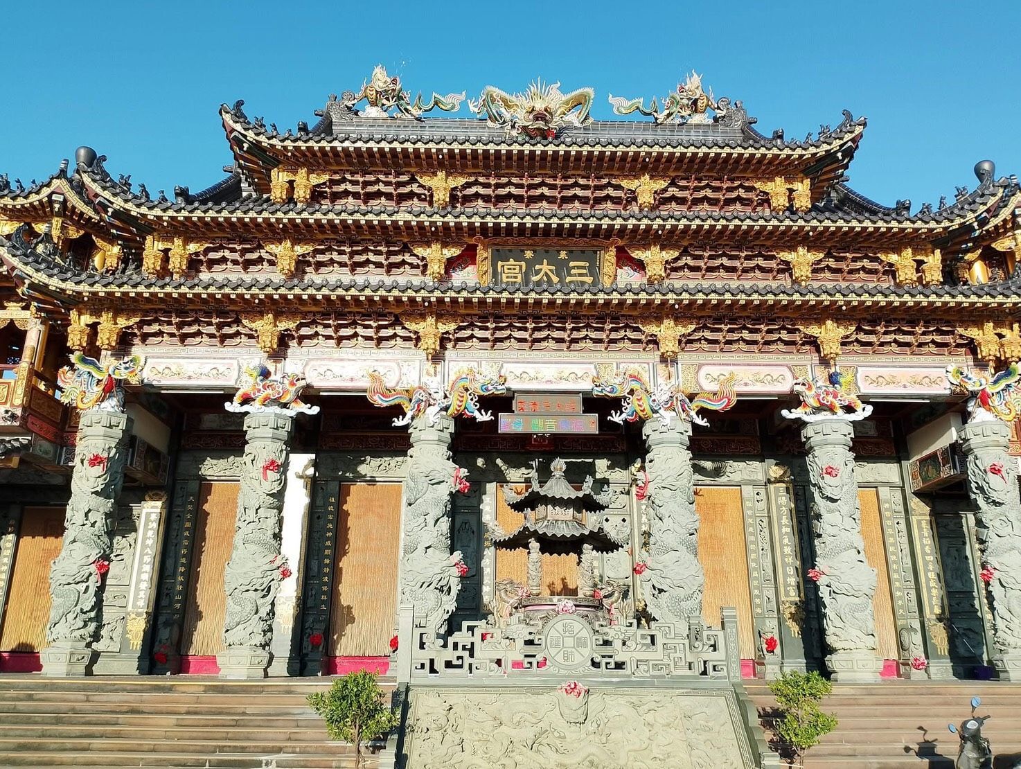 華麗的中國寺廟正面，雕刻精美的柱子，色彩斑斕的屋頂細節，以及藍天下懸掛的紅色燈籠。