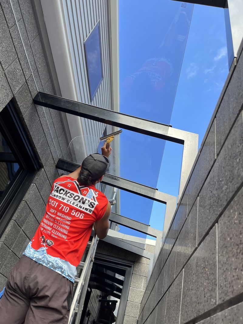 Person on Ladder Cleaning Glass Roof Panels Against a Bright Blue Sky — Jackson's Window Cleaning in Nerang, QLD