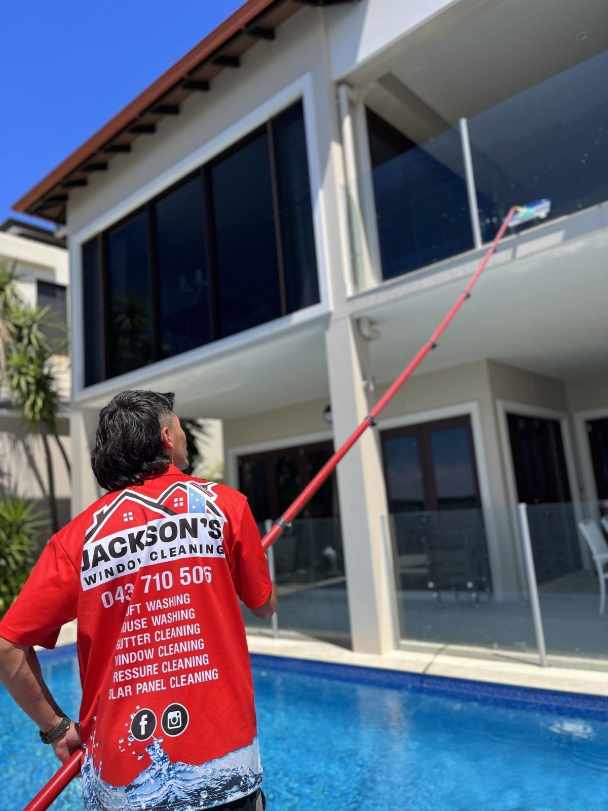 Man in red shirt cleans window of a modern house, using a long-handled brush, by a pool. — Jackson's Window Cleaning in Nerang, QLD