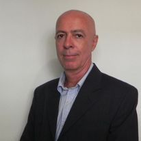 Um homem careca de terno e camisa listrada está parado em frente a uma parede branca.