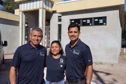 Three people in dark shirts stand smiling in front of a modern home under construction.