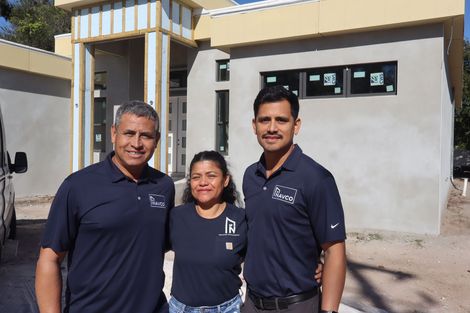 Three people in dark shirts stand smiling in front of a modern home under construction.