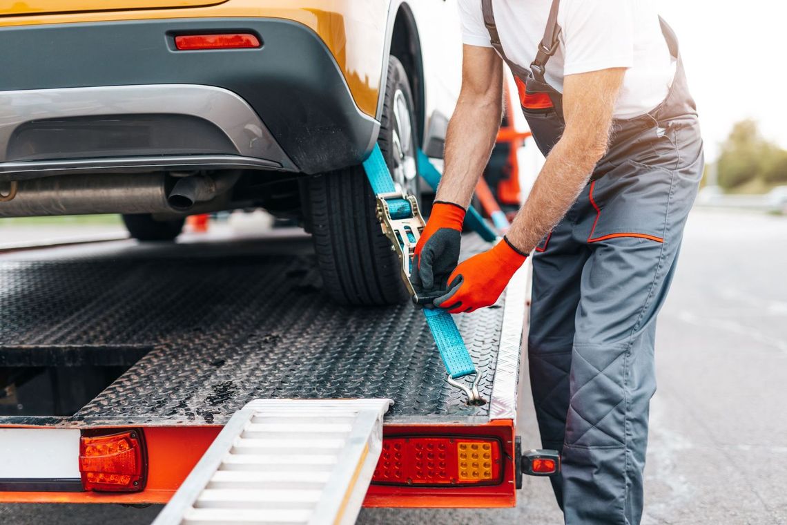 Tow truck operator securing a car on a flatbed with a blue strap and red gloves