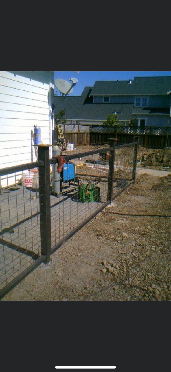 A partially built black metal fence in a yard with construction equipment visible. Houses and blue sky in background.