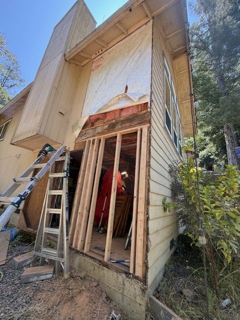 Construction site: House exterior being renovated. Exposed framing, siding, tar paper, and a person working inside.