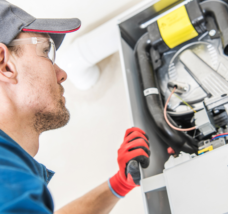 A worker in red gloves inspecting a furnace. He wears safety glasses, a cap, and blue coveralls.
