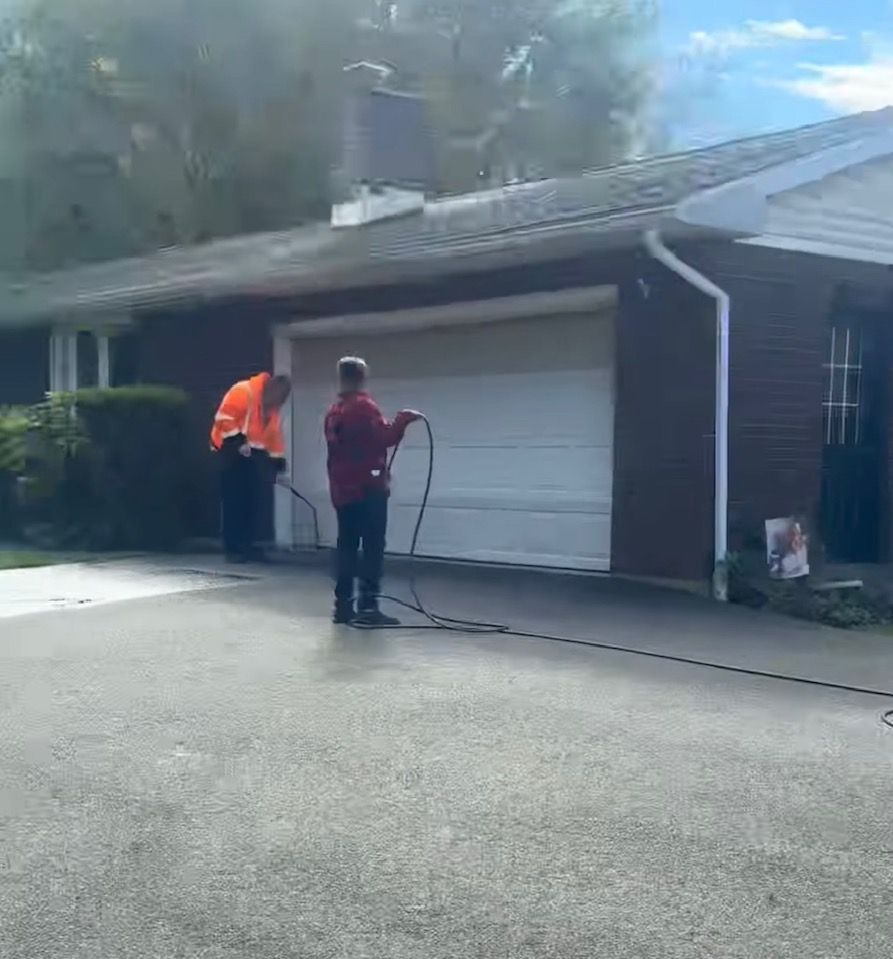 Two people in work clothes spray-wash a driveway in front of a brick house with a white garage door.