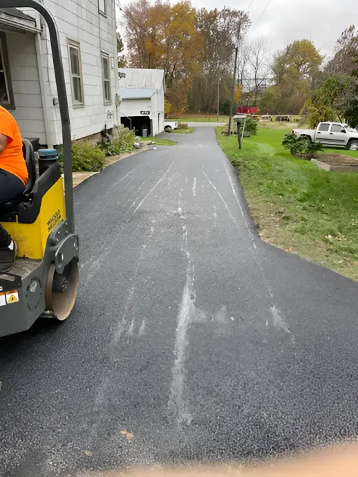 A bright yellow asphalt roller sits on a freshly paved driveway leading to a house, with a white truck parked nearby.