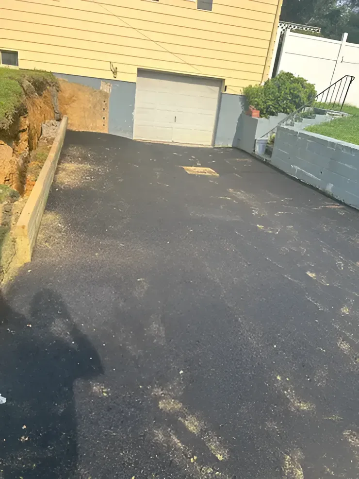 A paved driveway leads to a garage, flanked by a dirt embankment on the left and a stone retaining wall on the right.