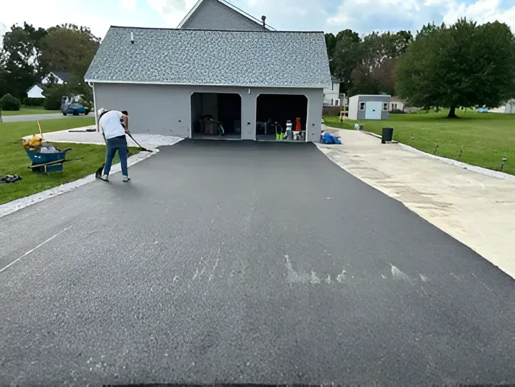 A worker uses a rake to smooth a freshly paved black asphalt driveway in front of a gray garage.