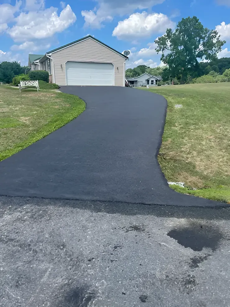 A newly paved, dark asphalt driveway leads to a suburban house with a white garage door on a sunny day.