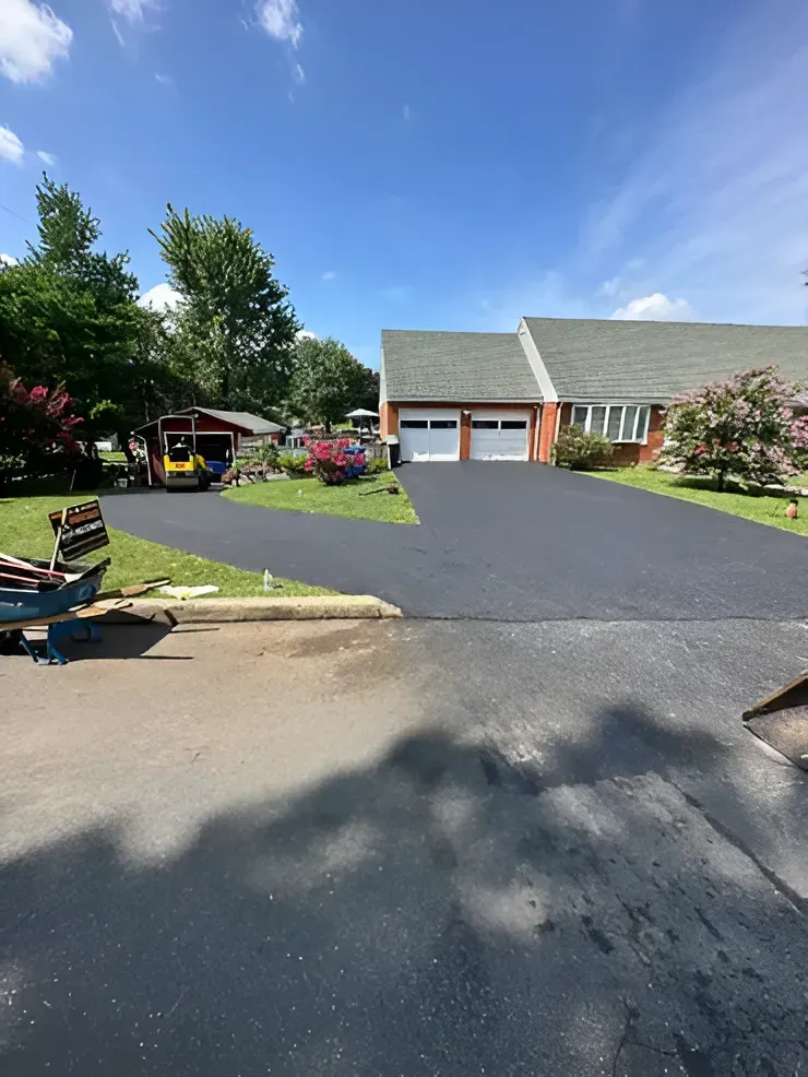 A freshly paved, dark asphalt driveway leading to a two-car garage attached to a suburban brick house on a sunny day.