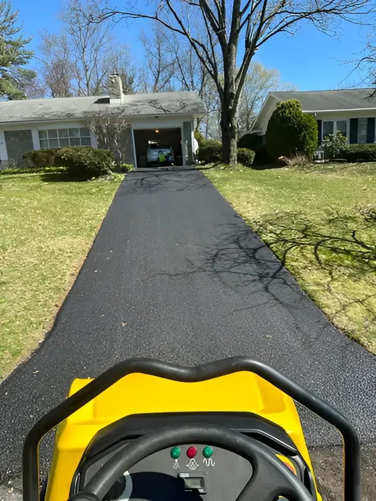 A view from the operator's seat of a yellow construction roller, facing a freshly paved asphalt driveway leading to a house.