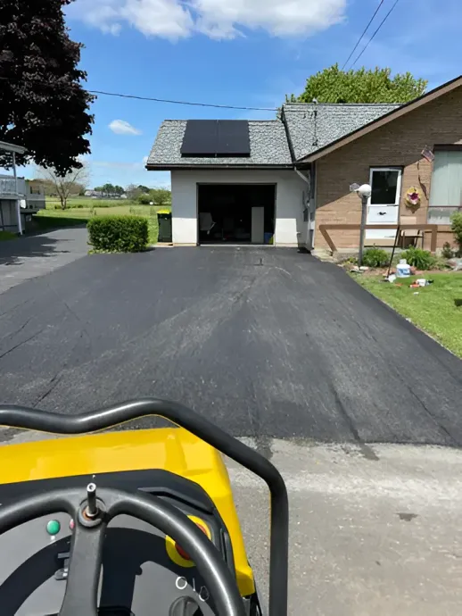 A view from the driver's seat of a yellow roller facing a newly paved asphalt driveway leading to a garage.