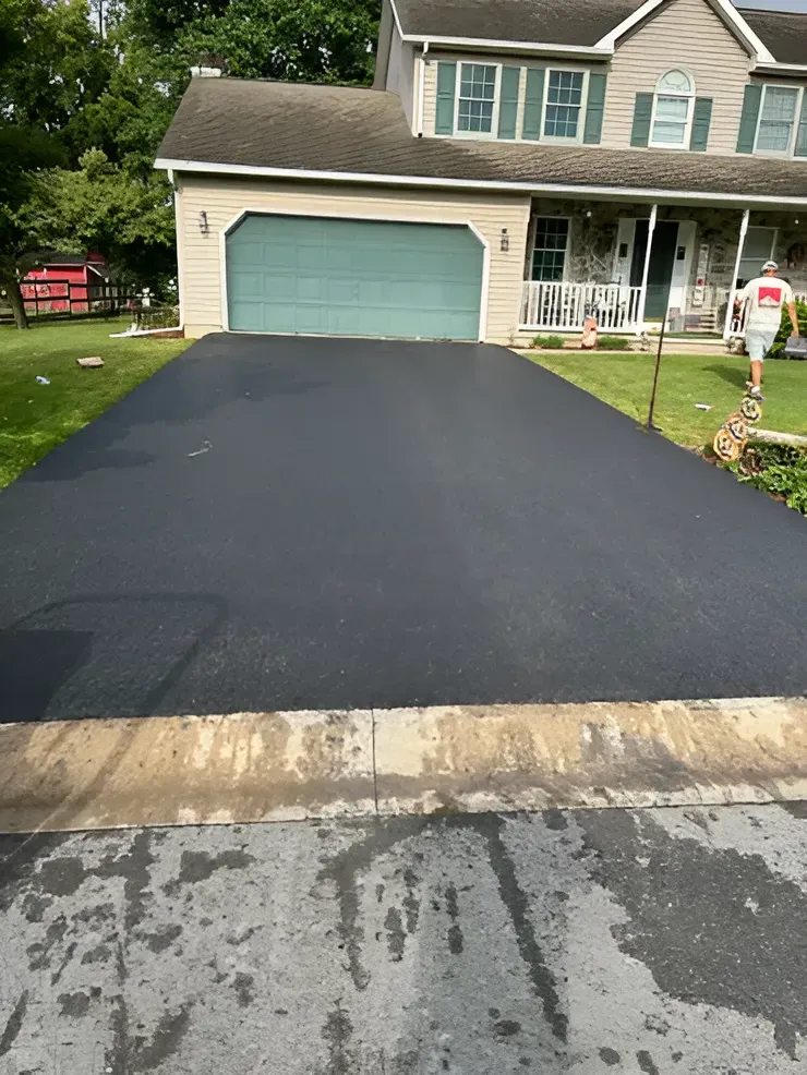 A freshly paved, dark asphalt driveway leading to a two-story tan house with a green garage door.