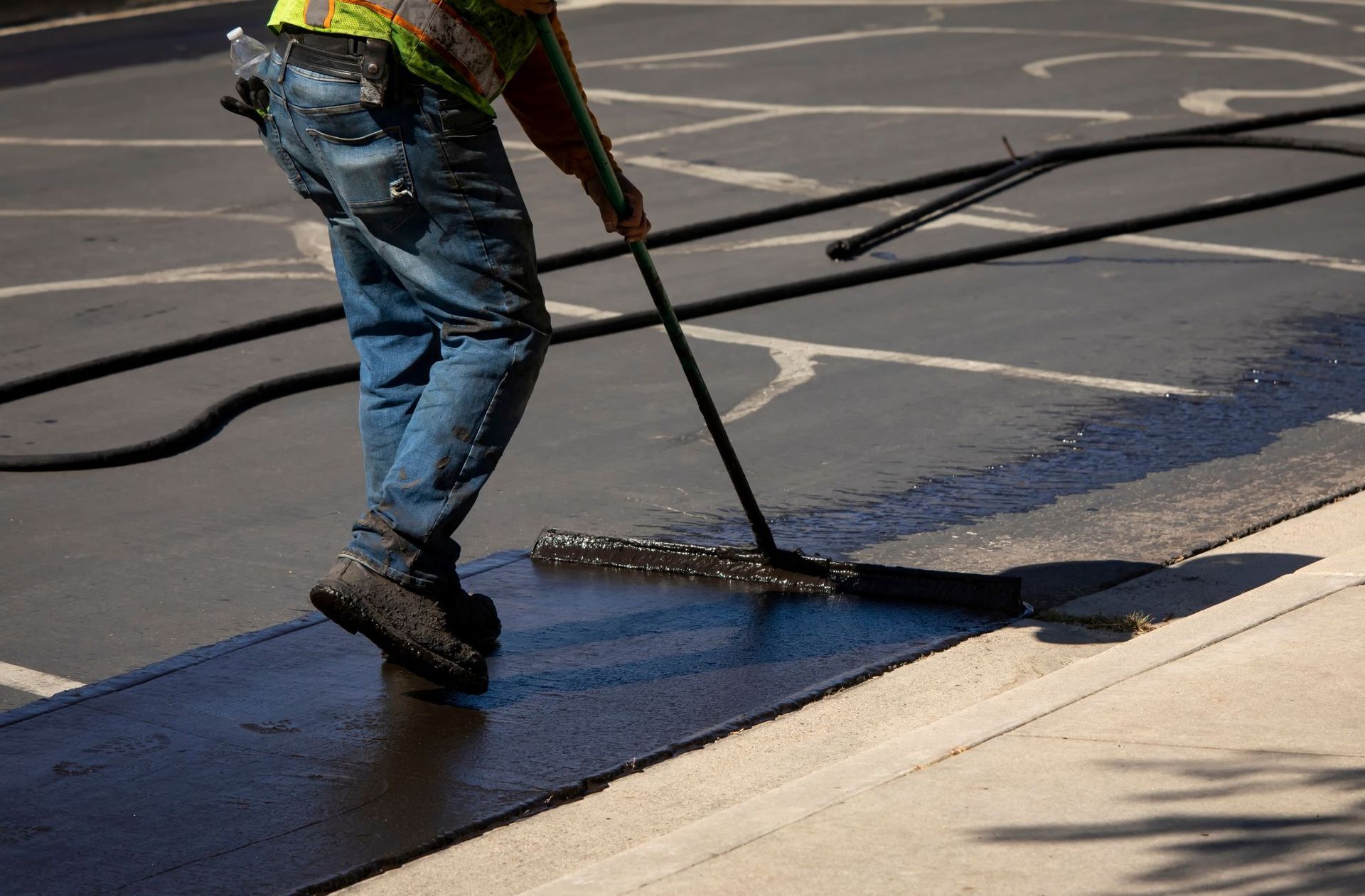 A worker uses a squeegee to spread black asphalt sealant across a paved surface.