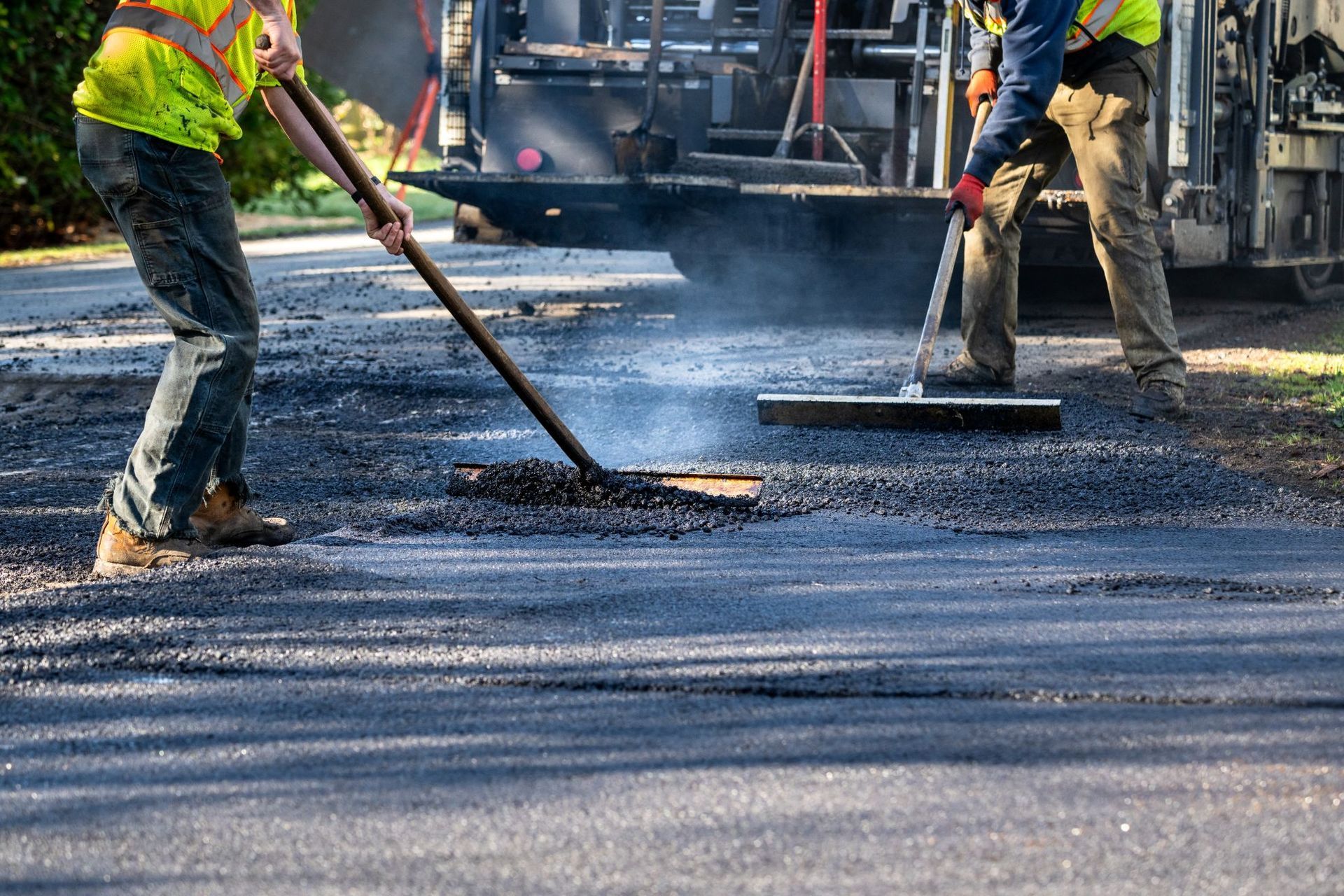 Two workers in high-visibility gear spread and smooth hot asphalt on a road.