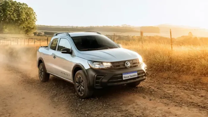 SUV prateado trafegando por uma estrada de terra poeirenta ao pôr do sol, com campos e árvores douradas ao fundo.