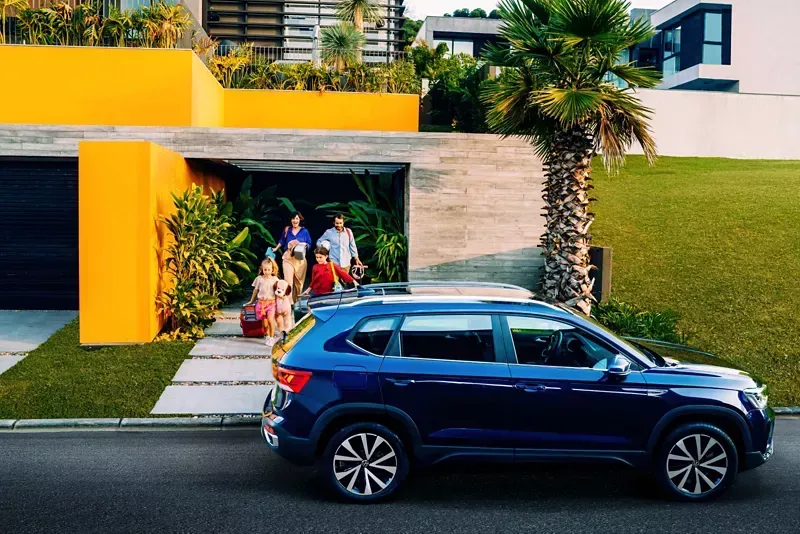 Um SUV azul está estacionado em frente a uma casa moderna; várias pessoas estão em um recuo na entrada, próximo a uma palmeira.