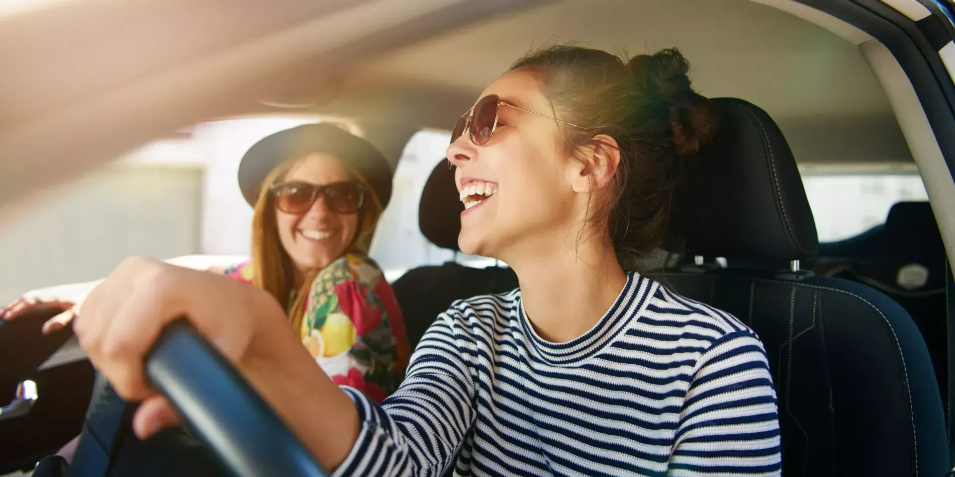 Duas pessoas sorridentes dentro de um carro, uma dirigindo e a outra de óculos escuros no banco do passageiro.