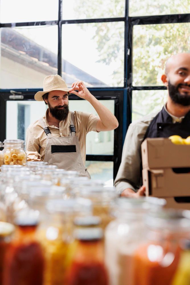 Market worker in a straw hat sorting jars on a table, with another person carrying boxes nearby.
