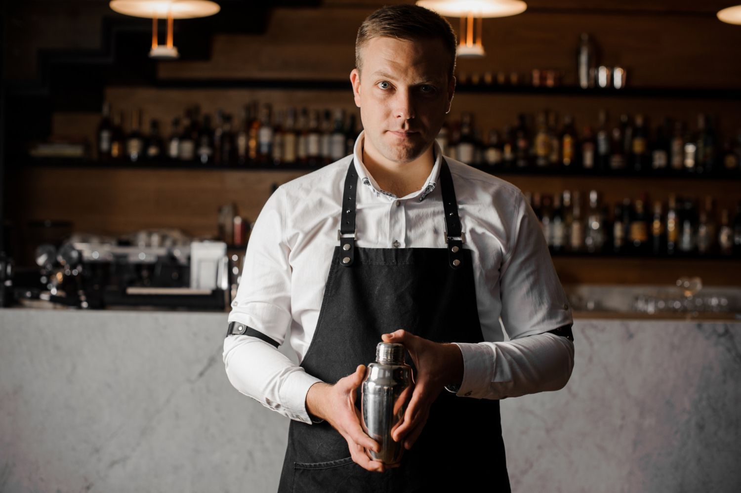 Bartender in white shirt and black apron holding a cocktail shaker behind a bar.