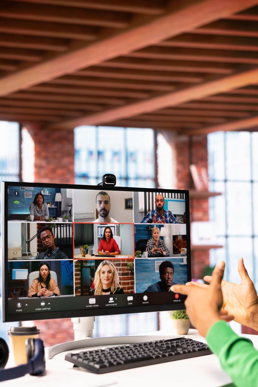 Video conference on a desktop monitor in a bright office, with a person gesturing near the screen.