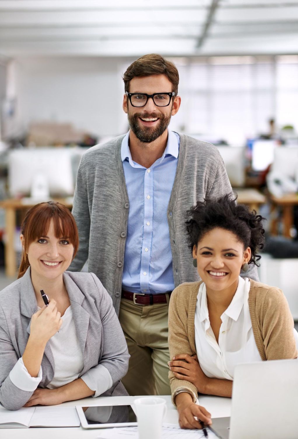 Three smiling coworkers in an office, seated at a desk with a laptop and tablet.
